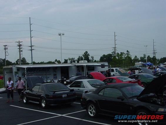 Several Ford Mustangs parked outside a trailer with hoods up. Some people are standing around