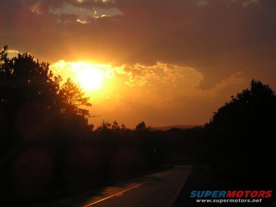 Sun setting behind clouds over a road with trees on the sides. Supermotors.net logo in corner