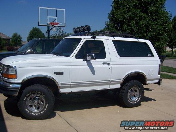 White Ford Bronco with roof rack, off-road lights, and chrome trim. Side view