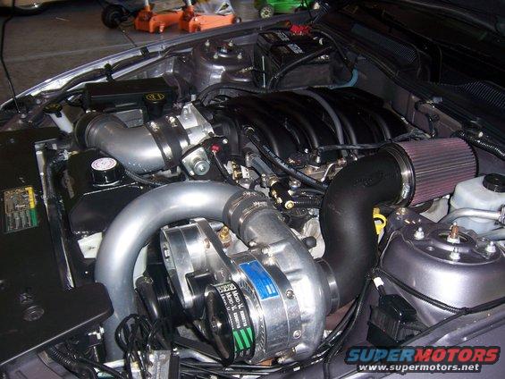 Engine bay with a polished supercharger, black intake piping, and a red air filter. The engine has black covers