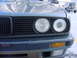1989 BMW 3 Series - Close-up of the front of a 1989 BMW 3 Series, showing the grille and headlights. An SUV is visible in the background.