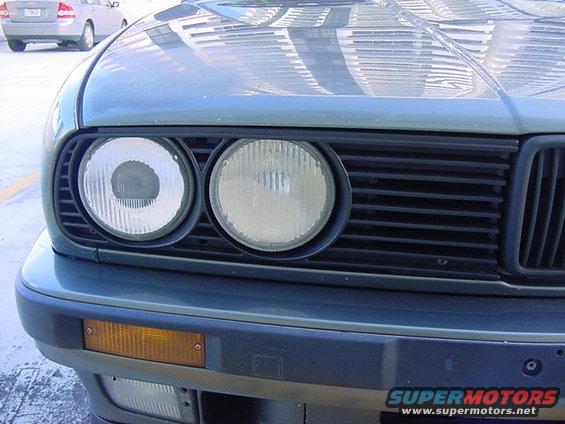 Front view of a 1989 BMW 3 Series with dual round headlights and a black grille. Another car is visible in the background