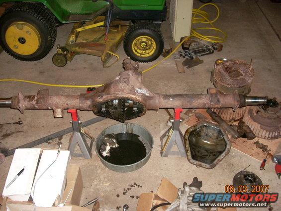Rusty differential axle assembly on jack stands with oil pan and brake drums nearby