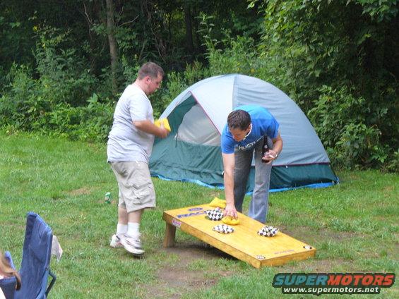 Two men playing cornhole in a grassy area with a tent behind them