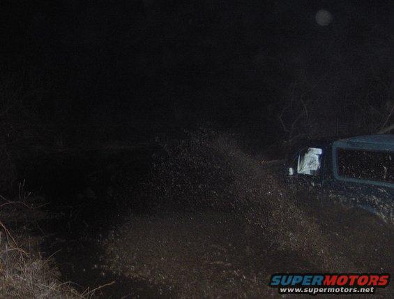 SUV driving through mud at night, kicking up spray. Watermark at bottom right