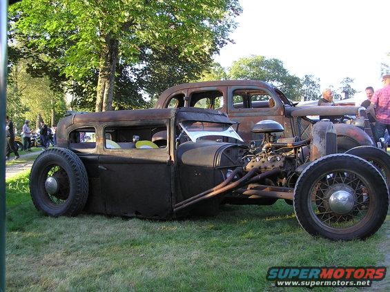 Rat rod with exposed engine and custom exhaust pipes, parked on grass