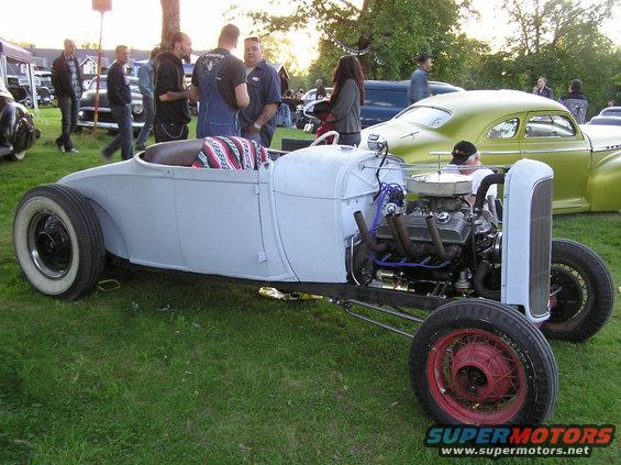 Custom hot rod roadster with exposed engine and whitewall tires parked on grass. Other cars and people are in the background