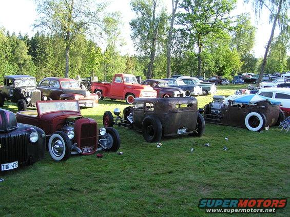 Several hot rod cars parked on a grassy field under trees