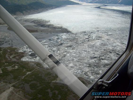 view of a glacier and ice chunks in water from an airplane window, with a wing strut in the foreground