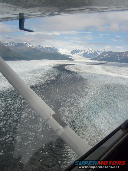 View from an aircraft window of a vast glacier flowing between snow-capped mountains under a cloudy sky