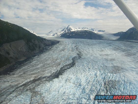 Aerial view of a vast glacier flowing between mountains under a cloudy sky. Part of an aircraft is visible in the upper right