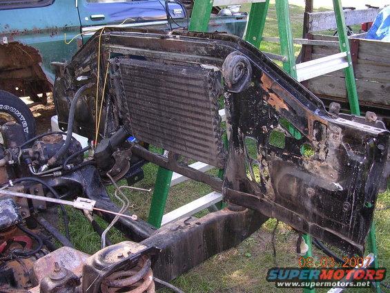 Disassembled front end of a vehicle, showing the radiator and frame. A blue truck is partially visible in the background