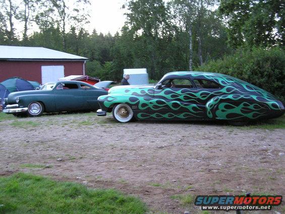 Two custom hot rod cars parked side-by-side, one dark green coupe and one black car with green flames