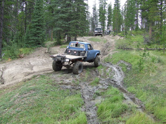 Blue pickup truck with flatbed driving up a muddy hill, another vehicle behind it