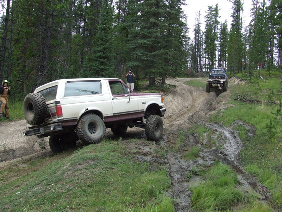 White and maroon 1990 Ford Bronco climbing a muddy hill, another blue off-road vehicle behind it
