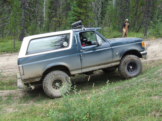 Dark green and white Ford Bronco with off-road tires on a dirt path, mud splattered on the lower body