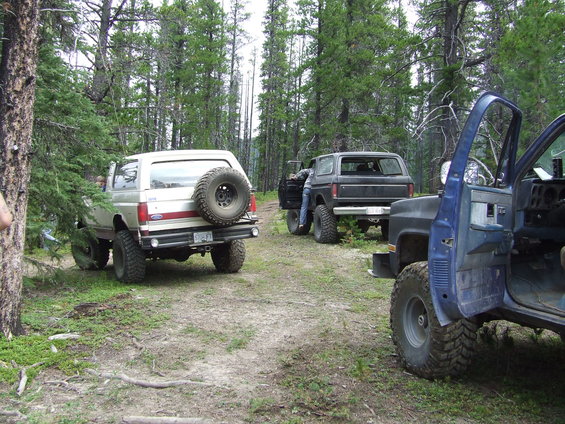 1990 Ford Bronco with spare tire on back, parked on dirt road in forest with two other SUVs