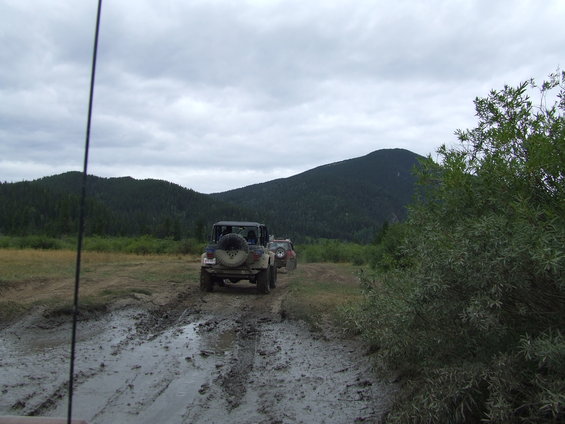 Several off-road vehicles drive through a muddy track with trees and a mountain in the background