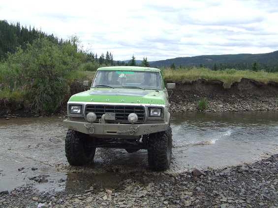 Green Ford Bronco with aftermarket bumper and large tires driving through a rocky stream
