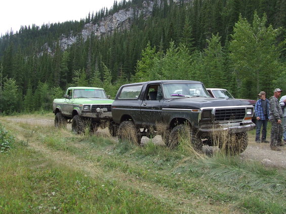 Black 1978 Ford Bronco and green Ford pickup truck parked on a dirt road with trees and a mountain in the background