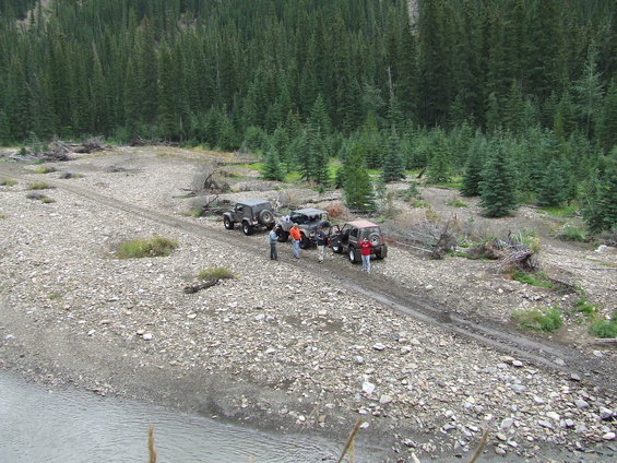 Several Jeeps parked on a rocky dirt road with people standing around them. Forested mountains are in the background