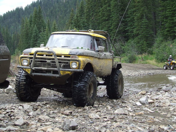 Yellow Ford pickup truck with large off-road tires and a brush guard, parked on a rocky trail. Another ATV is visible in the background