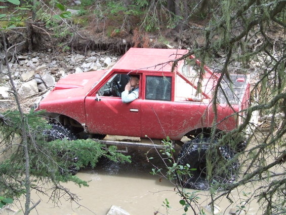 Red off-road vehicle with large tires driving through muddy water, driver visible inside
