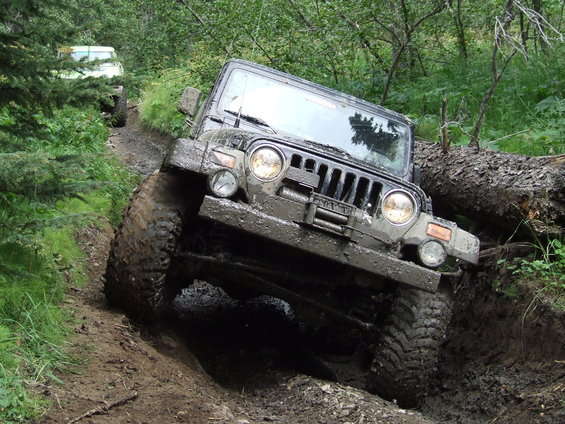 Muddy Jeep Wrangler climbing a steep, wet trail with a fallen tree to the right. Another white SUV is visible further up the path
