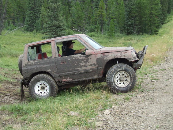 Mud-covered Suzuki Samurai driving off-road on a dirt path with trees in the background