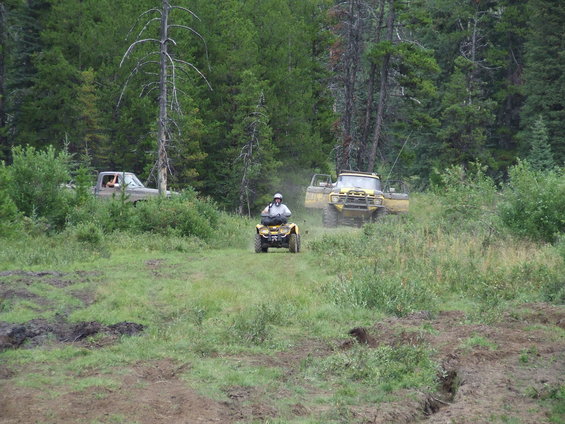 Person on yellow ATV riding toward a yellow off-road truck with doors open. Another truck is partially visible behind trees
