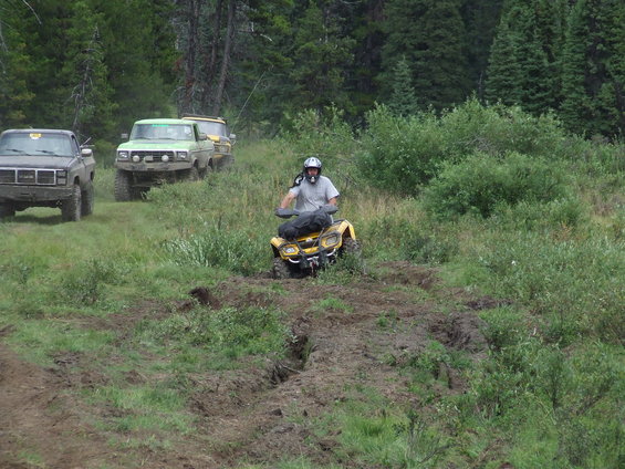 Yellow ATV with rider on a muddy trail, with trucks in the background