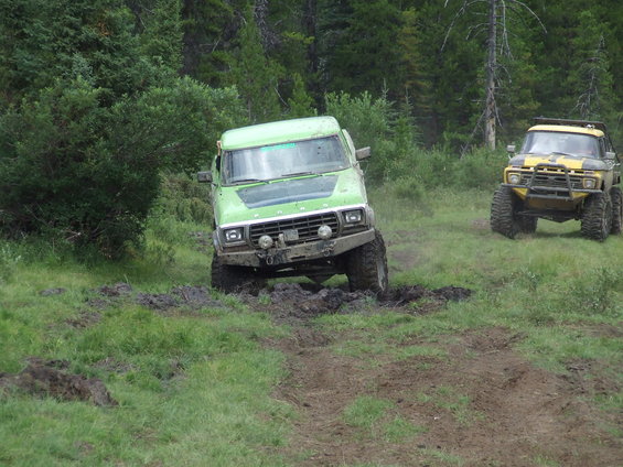 Green off-road truck stuck in mud, yellow truck behind it on grassy trail