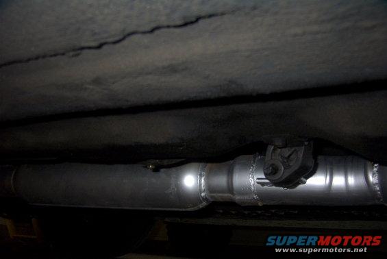 Close-up of a vehicle's exhaust pipe and hanger, with a SuperMotors.net watermark