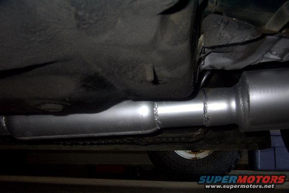 Underside of a vehicle showing a new exhaust pipe and muffler. A tire is visible