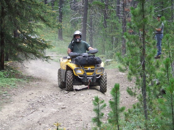 Yellow ATV with a rider wearing a helmet on a dirt road in a forest. Another person stands on the side of the road