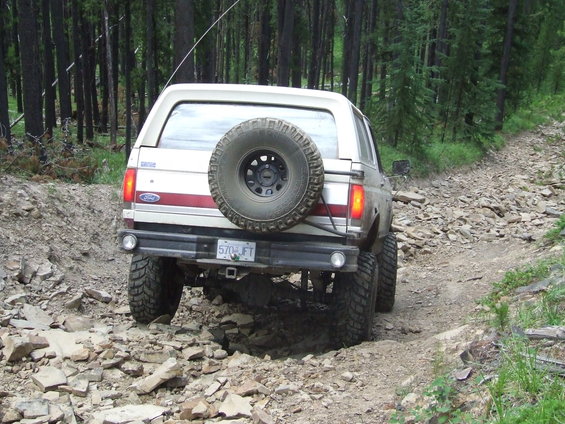 Rear view of a 1990 Ford Bronco driving on a rocky, wooded trail. It has a spare tire mounted on the back and a license plate reading 570 JFT