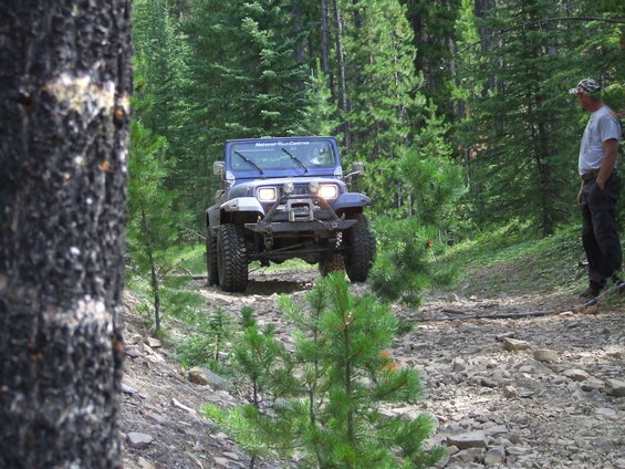 Blue Jeep Wrangler driving on a rocky, wooded trail. A person stands on the side of the trail watching