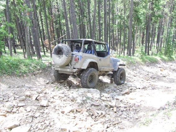 Jeep Wrangler TJ with spare tire on back, driving on a rocky forest trail