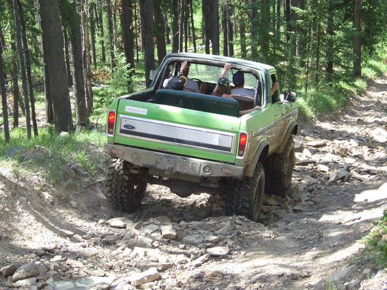 Green 1978 Ford Bronco with people inside driving on a rocky forest trail