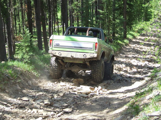 Green Ford Bronco driving on a rocky forest trail. Driver and passenger have arms raised