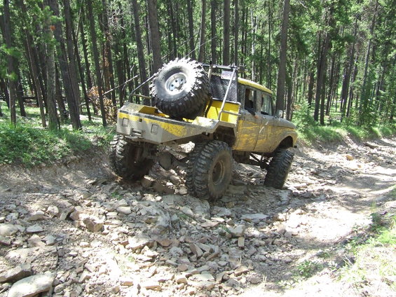 Yellow custom off-road truck with large tires and spare tire on back, driving on a rocky forest trail