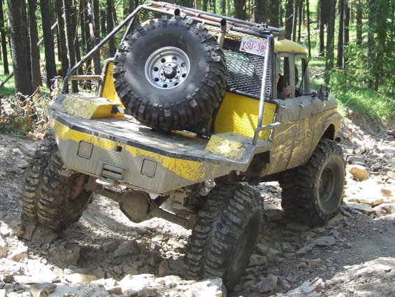 Yellow modified 1966 Ford Bronco with large tires and a spare tire on the back, driving on a rocky trail