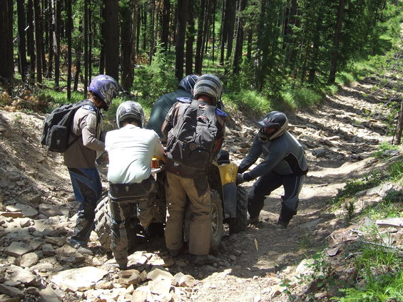 Four people in helmets pushing a yellow ATV stuck on a rocky forest trail