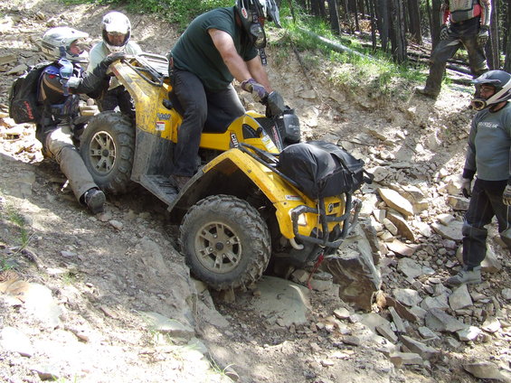 Yellow ATV with three riders navigating a steep, rocky incline, one rider is being pushed
