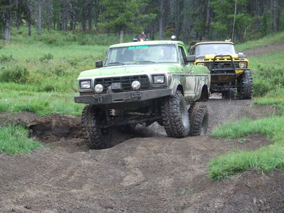 Green lifted Ford F-Series truck with custom bumper, yellow lifted Ford F-Series truck behind it, on muddy trail