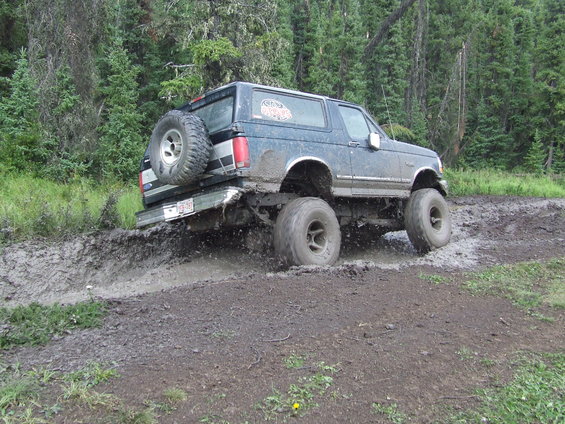 Blue 1990 Ford Bronco driving through mud with large tires and spare on back