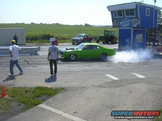 Green muscle car doing a burnout on a drag strip, with a tractor and pickup truck in the background