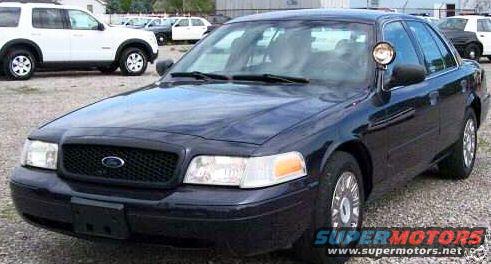 2003 Ford Crown Victoria police car with spotlight on driver's side mirror, parked in gravel lot with white SUV and other cars