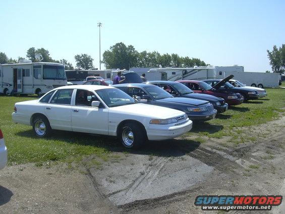 White sedan parked on grass, side view, chrome wheels, some tire marks on dirt