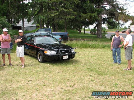 Black Ford Crown Victoria with a push bar, parked on grass with people around. License plate reads 569-GUA